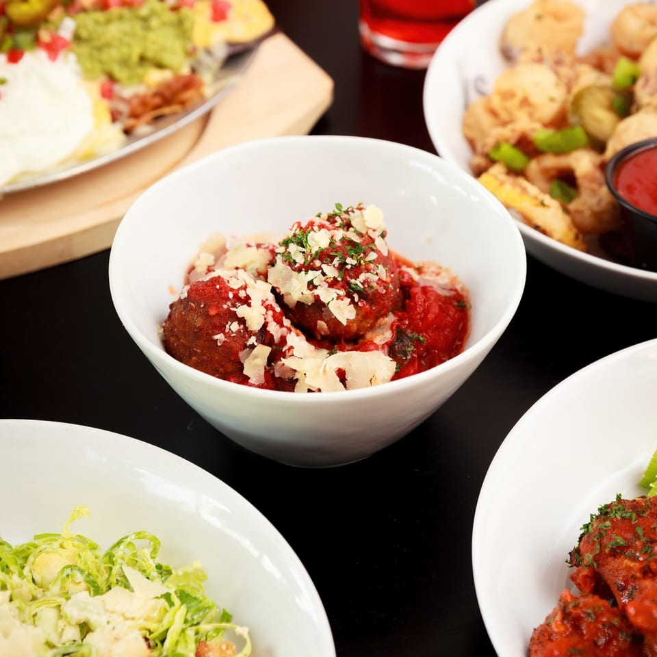 A white bowl of meatballs with red sauce and parmesan cheese, surrounded by other dishes.