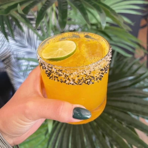 Hand holding a vibrant orange cocktail with a salted rim and lime slice, against a leafy green background.