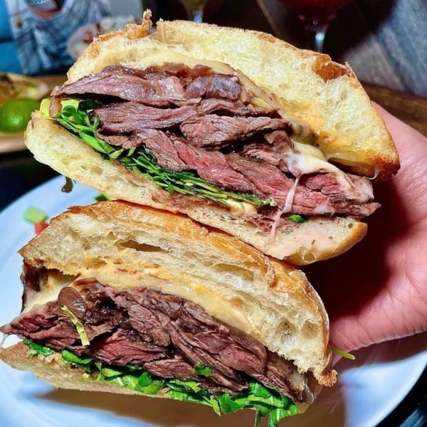 A hand holds a sliced Salted. On The Harbor steak sandwich with melted cheese and greens.