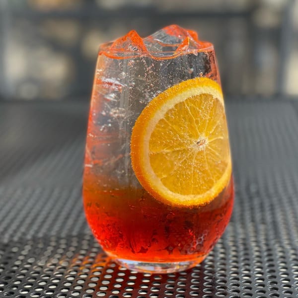 A vibrant red cocktail with ice and an orange slice, on a dark, perforated table.